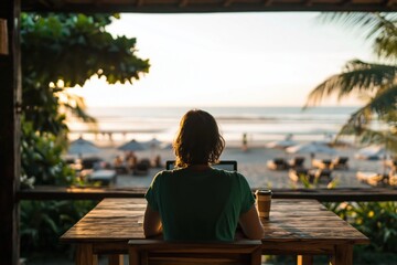 Obraz premium Person sits at wooden table with laptop, facing ocean at tropical beach during beautiful sunset. High quality photo