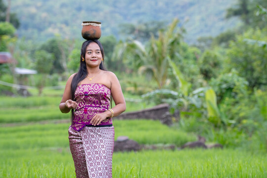 Balinese woman balancing basket on head in traditional dress in rice field.