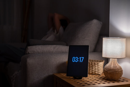 Person lying awake in couch in a dark room during the early hours, experiencing insomnia with a digital alarm clock displaying 03:17 am on the bedside table