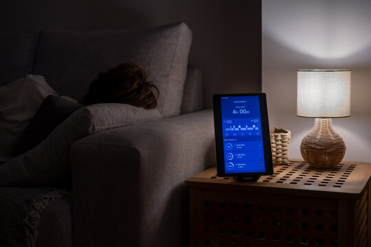 Person lying on a couch in a dark room, attempting to sleep while a tablet on a nightstand displays fragmented sleep data, indicating insufficient rest and the impact of technology on sleep health