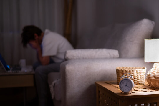 Exhausted woman struggling with sleep deprivation and stress, sitting in a dark room with a laptop while an alarm clock shows the early morning hours, indicating insufficient rest