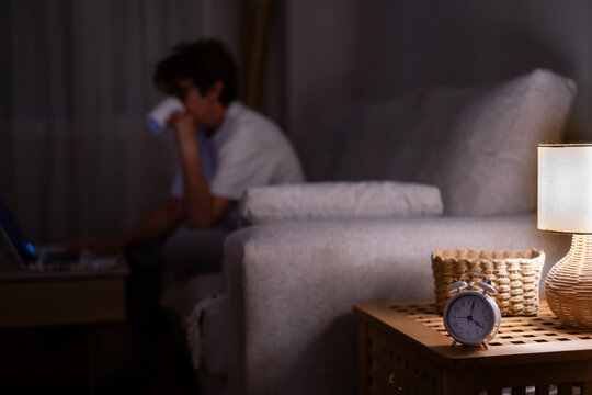 Young woman battling insomnia and exhaustion at a home desk, working on a laptop late into the night with coffee and an alarm clock showing early hours, dim lamp glow