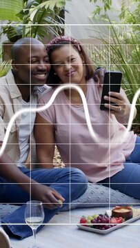 Vertical video: Couple sitting in garden woman angling phone for social selfie, squiggle over phone