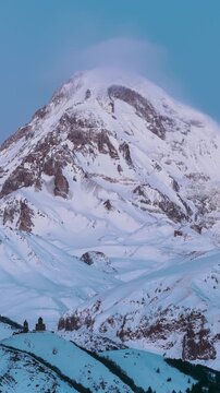 Stepantsminda, Gergeti, Georgia. Peak Of Mount Kazbek Covered Snow And Famous Gergeti Trinity Tsminda Sameba Church In Winter Landscape. Beautiful Georgian Winter Nature Landscape