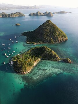 Aerial view of Flores islands featuring lush green hills, turquoise waters, coral reefs, and anchored boats in Labuan Bajo, East Nusa Tenggara, Indonesia.