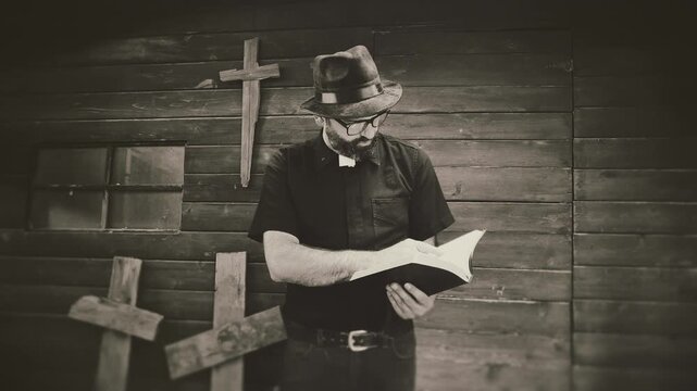 Clergyman, reverend or priest wearing a clerical collar and reading a bible. Preacher preaching the gospel in front of an old rustic rural church. Edited in vintage film style black and white.