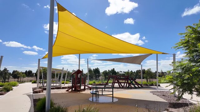 A vibrant yellow triangular shade sail stretches over a modern children's playground in a well-maintained Australian suburban park. Sun protection,&nbsp;outdoor recreation infrastructure.