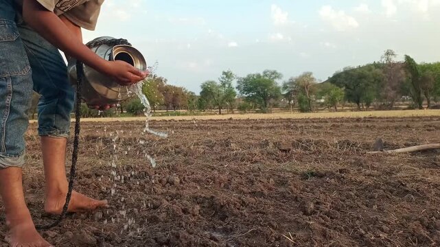 Side view of an Indian boy pouring water from a traditional steel pot onto dry soil in a rural field under a bright sky