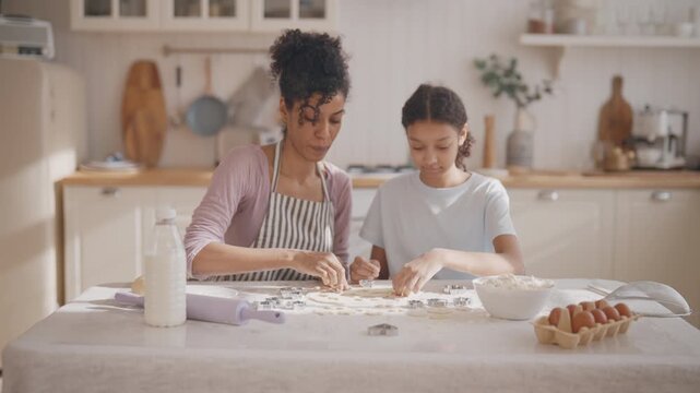 Master class for teen girl how to make homemade cookies, african woman and girl. Housewife teaching her daughter to cook and bake, happy smiling people in cozy home kitchen interior, medium shot