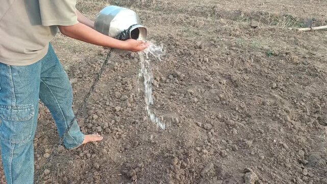 Village boy pouring water from a steel vessel onto dry farmland with traditional farming tools in the background