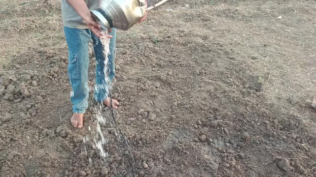 Village boy pouring water from a steel vessel onto dry farmland with traditional farming tools in the background
