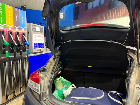 Maleta en maletero abierto de coche en gasolinera con mangueras de surtidor para repostar en viaje de vacaciones y escapada en plan ocio 