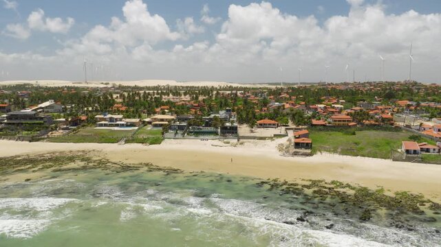 Ta&iacute;ba village S&atilde;o Gon&ccedil;alo do Amarante Cear&aacute; Brazil with beachfront houses, reef exposed shoreline, tidal pools and wind turbines over dune field, drone descent establishing shot