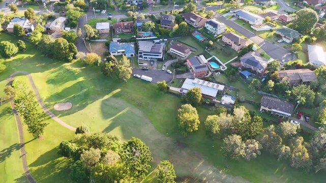 Aerial view of suburban houses bordering lush green golf course fairway during golden hour.