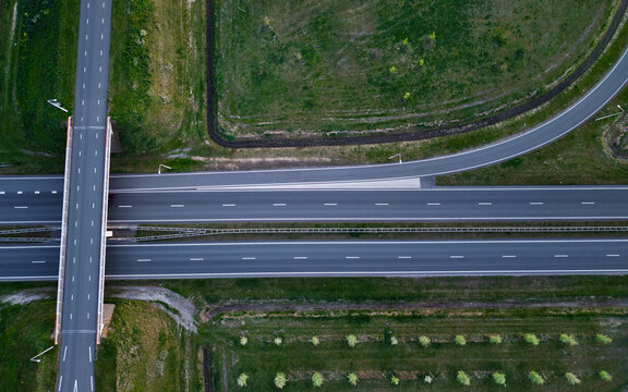 Aerial view of a multi-lane highway interchange featuring a bridge overpass, lush green fields, and organized rows of trees.