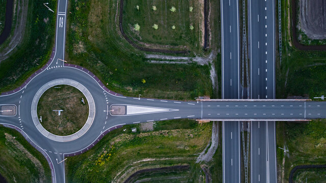 Aerial view of a highway interchange featuring a roundabout and a bridge over a multi-lane motorway surrounded by green fields and trees.