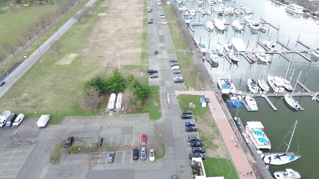 Waterfront parking zone lines Audrey Zapp Dr beside Liberty Landing Marina, Morris Canal Basin with open lawns and service tanks marking park edge. Pavement grids, boat hulls, soft gray light, NJ
