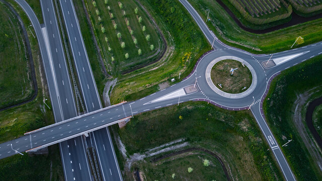Aerial view of a modern highway interchange featuring a circular roundabout and a bridge over a multi-lane road surrounded by lush green fields and rows of trees.