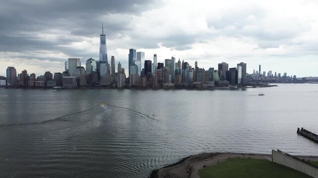 Harbor to skyline span views Hudson River from Liberty State Park toward Lower Manhattan with clustered towers rising behind waterway. Rippling surfaces, vessel trails, soft gray cloud layers, NJ