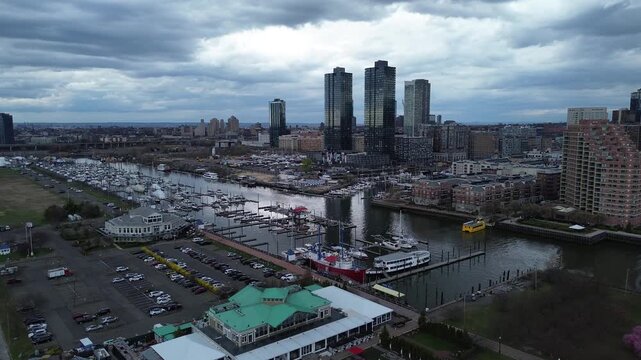Marina basin overview captures Liberty Landing Marina along Audrey Zapp Dr with slips, piers, Morris Canal Basin curving Hudson. Mixed hull, asphalt lots, soft overcast tones subdued contrast, NJ