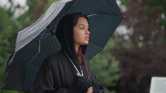 Young woman under umbrella standing stoic, rain streaking umbrella surface, hood pulled over head, urban park lanes blurred in background, commuter posture with necklace visible, calm resilient mood