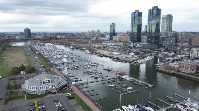 Harbor district density spans Liberty Landing Marina, waterfront blocks near Morris Canal Basin with mid rise, high rise forms rising inland. Vessel lines, roof textures, soft atmospheric haze, NJ