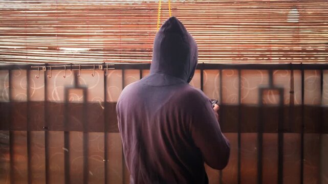 Man in black hoodie behind curtain using phone to type messages or send secret email. Mysterious person indoors holding smartphone. Hacker or spy in a house.