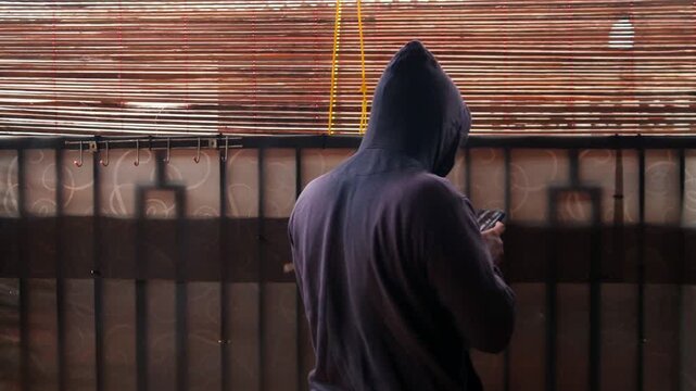 Man in black hoodie behind curtain using phone to type messages or send secret email. Mysterious person indoors holding smartphone. Hacker or spy in a house.