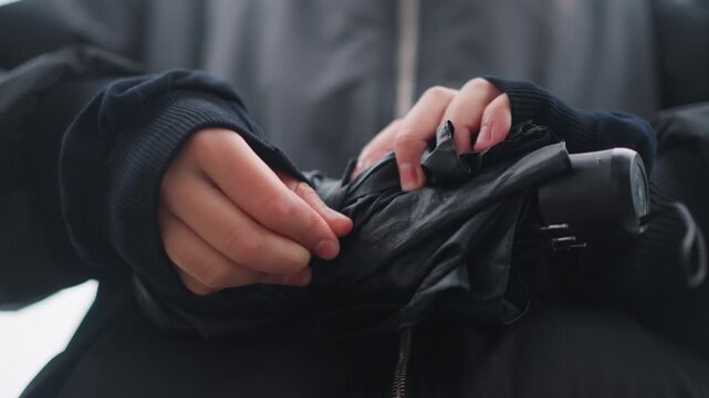 Woman methodically closing soaked umbrella slow fingers compress compact frame, black jacket zipper visible, raindrops glistening, quiet urban pause, careful tactile focus on fabric and ribs