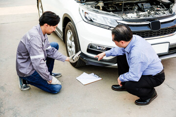 Fototapeta premium Mechanic and client kneel by a white car with the hood open, inspecting front bumper damage and discussing repairs beside a clipboard.