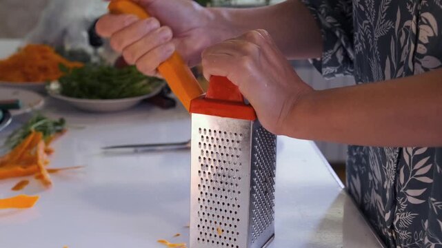 woman grating carrot side view shredding vegetable on metal grater over kitchen surface, preparing fresh ingredients for cooking meal, healthy eating routine, home food prep process