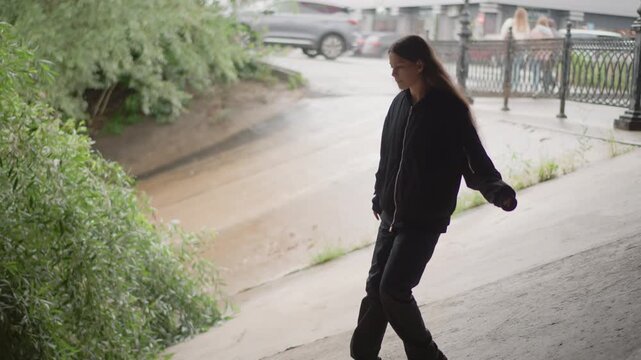 Young woman rain soaked path walking, urban explorer moves along iron railing and lamp posts, wet pavement and puddles, green embankment and sandy bank nearby, cautious step toward river vegetation,