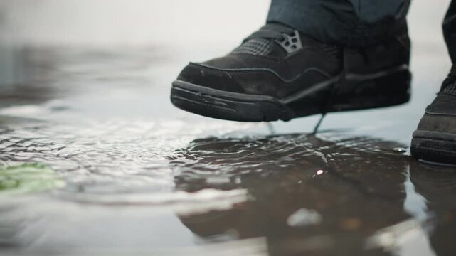 Woman stepping into puddle, wet shoes and reflection. Closeup of black boots splashing on city sidewalk with shallow reflections, laces loose, muted gray light, slowmotion feel, commuter vibe turning