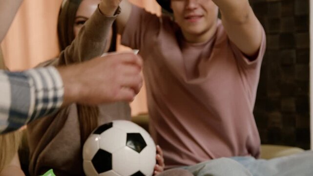 Group of young adults celebrating a sports event at home, holding drinks and a soccer ball, with excitement visible in their expressions and gestures