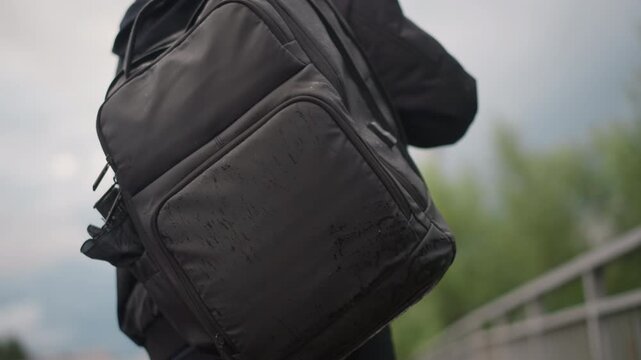 Woman walking across bridge in rain with large backpack, rainclouds and wet railing frame cityscape, steady stride and focused posture show commuter determination and travel intent