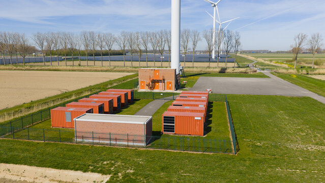 Aerial view of a battery energy storage system with orange containers and wind turbines under a clear blue sky in Middelharnis, Zuid-Holland, Netherlands.