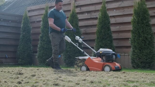 A man uses a professional gasoline scarifier to remove moss and thatch from a green lawn in a private house backyard.