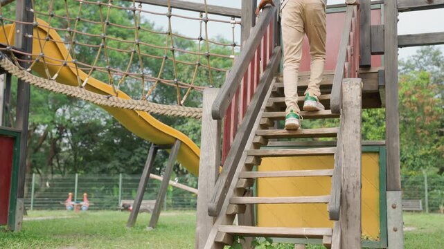 Young son climbing playground wooden stairs, ascending toward slide, grass underfoot, worn timber steps, tentative balance, sunny park setting, small sneakers, focused curiosity, learning milestone