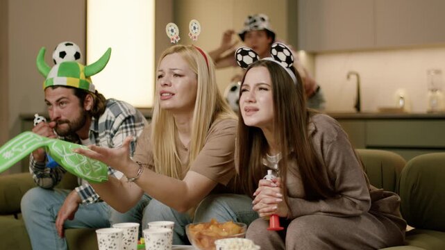 Group of friends passionately watching football match at home, wearing playful hats and holding cheering props, surrounded by snacks and drinks on the couch