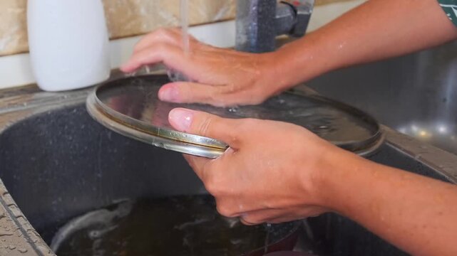 Woman washing pan lid at kitchen sink in side view, rinsing grease and soap under running water, cleaning cookware after cooking, daily household routine and kitchen hygiene process