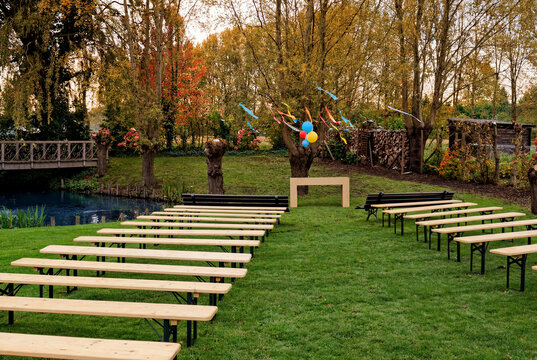 Outdoor garden ceremony setup with wooden benches on green lawn. Rustic wedding venue by pond with balloons and autumn trees. Festive event decoration in nature.