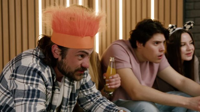 Group of friends passionately watching football match at home, man in orange hat leans forward, while others react with excitement and anticipation in a cozy living room setting