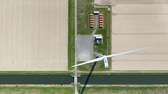 Aerial view of a large white wind turbine standing over a canal with a small bridge, containers and geometric agricultural fields in Middelharnis, Zuid-Holland, Netherlands.