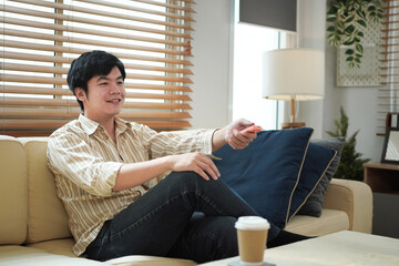 Fototapeta premium young asian man sitting on couch change channel with remote controller watching TV relaxing at home, male choose movie television program at home resting
