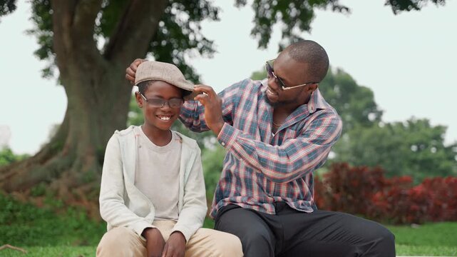 Black father and son beneath tree sharing playful grooming and warm laughter father adjusts sons headband and smooths hair while both sit on grass in casual clothes, sunlit park backdrop, tree