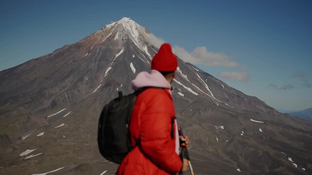 Portrait young man in red jacket and red knitted hat with large sunglasses on face stands against backdrop of majestic volcano with snow-capped peak. Slow motion