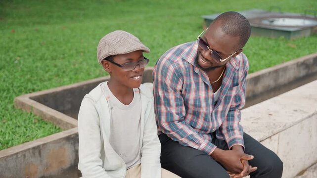 Green lawn black father and son laughing and exchanging advice on concrete ledge, father leaning in with warm smile while son grins in cap and glasses, relaxed friendly interaction in bright park