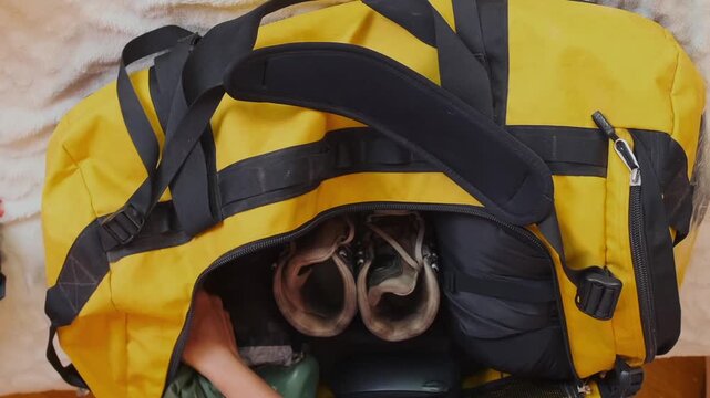 Overhead view of woman placing rain jacket and outdoor clothing into travel bag, preparing wardrobe for mountain hike, organizing apparel for camping, trekking and unpredictable weather conditions