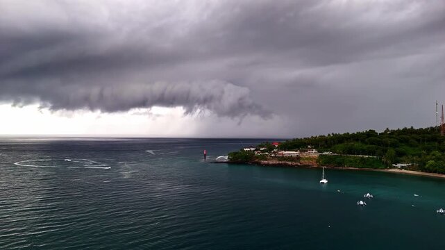 An ominous shelf cloud storm system moves over a tropical harbor with moored sailboats and lush green headland visible against turquoise calm waters. Intense weather contrast. Senggigi Beach