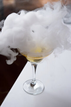 Smoking Cocktail in Coupe Glass with Dry Ice Effect. A close-up of a golden cocktail in a glass surrounded by thick white smoke or vapor from dry ice. Professional molecular mixology at a bar